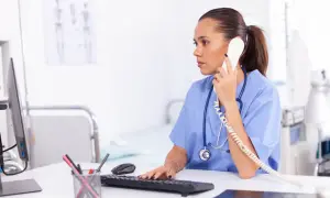 Rejestratorka medyczna w niebieskim uniformie rozmawia przez telefon, pracując przy komputerze w gabinecie.