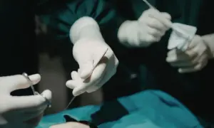 Dłonie w rękawiczkach chirurgicznych trzymają narzędzia. W tle widać anestezjologa czuwającego nad pacjentem.