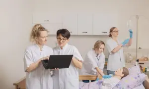 Pielęgniarka w białym kitlu uczy się obsługi laptopa, podczas gdy koleżanka ćwiczy zakładanie kroplówki przy fantomie.