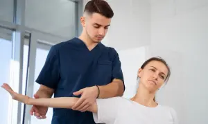 Fizjoterapeuta w granatowym uniformie delikatnie naciska na ramię pacjentki podczas ćwiczeń. Czy fizjoterapeuta to lekarz? Pomaga w rehabilitacji.