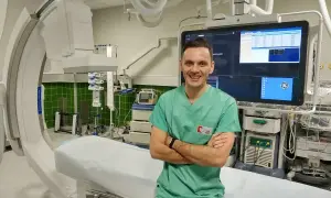 Uśmiechnięty technik elektroradiolog w zielonym fartuchu stoi na tle nowoczesnego sprzętu medycznego w sali zabiegowej.