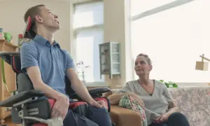 Młody mężczyzna z ALS w wózku inwalidzkim, z uśmiechem na twarzy, siedzi obok kobiety na kanapie.
