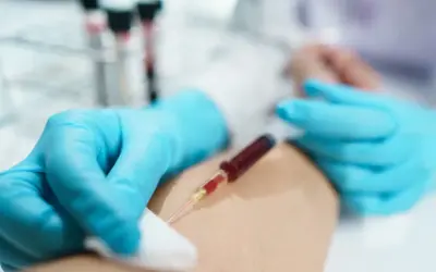 Felczer w niebieskich rękawiczkach pobiera krew do strzykawki z ręki pacjenta. W tle probówki.