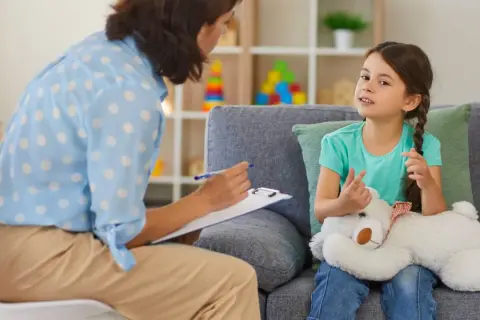 Psychoterapeutka z notatkami rozmawia z dziewczynką trzymającą misia.