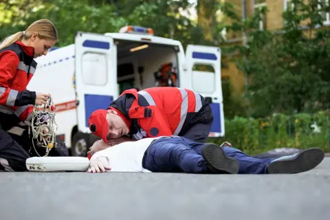 Ratownicy medyczni udzielają pomocy osobie nieprzytomnej na drodze, obok otwartej karetki.