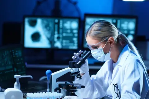 Diagnosta laboratoryjny w okularach i masce pochyla się nad mikroskopem, analizując próbki.