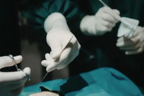 Dłonie w rękawiczkach chirurgicznych trzymają narzędzia. W tle widać anestezjologa czuwającego nad pacjentem.