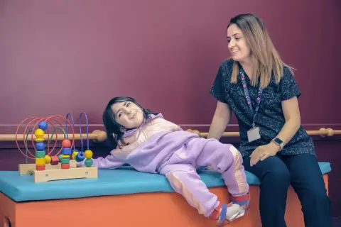 Uśmiechnięta dziewczynka w fioletowym dresie leży obok terapeuty, bawiąc się zabawką z koralikami.