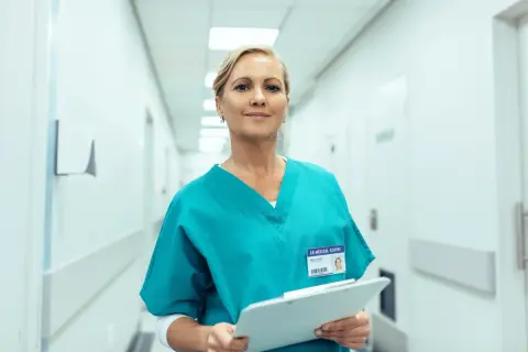 Uśmiechnięta farmaceutka w turkusowym uniformie trzyma dokumenty. Zastanawiasz się, ile zarabia farmaceuta?