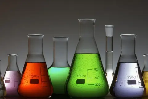 Kolorowe płyny w kolbach laboratoryjnych, od białego po żółty, zielony, pomarańczowy i fioletowy. Jedna z nich zawiera substancję przypominającą arszenik.