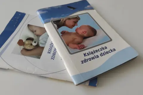 Dwie książeczki zdrowia dziecka leżą na białym tle. Jedna z nich przedstawia matkę całującą niemowlę, druga śpiące dziecko z misiem.