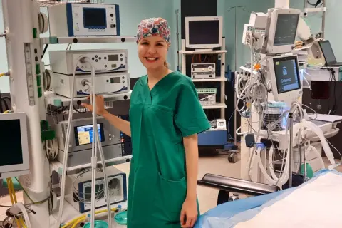 Uśmiechnięta instrumentariuszka w zielonym uniformie stoi obok aparatury medycznej na sali operacyjnej.