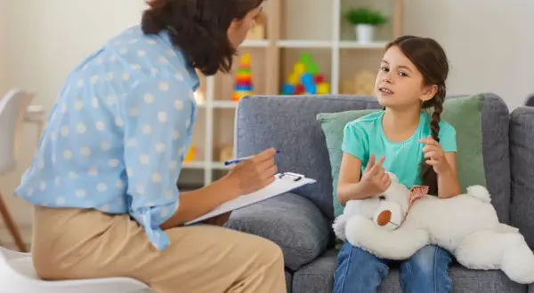 Psychoterapeutka z notatkami rozmawia z dziewczynką trzymającą misia.