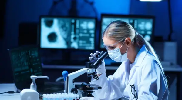Diagnosta laboratoryjny w okularach i masce pochyla się nad mikroskopem, analizując próbki.