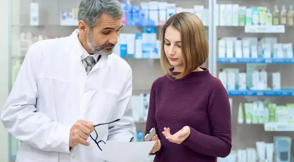 Doświadczony farmaceuta doradza klientce w aptece, wskazując na receptę i opakowanie leku.