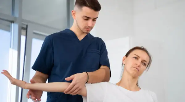 Fizjoterapeuta w granatowym uniformie delikatnie naciska na ramię pacjentki podczas ćwiczeń. Czy fizjoterapeuta to lekarz? Pomaga w rehabilitacji.