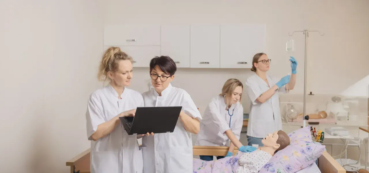 Pielęgniarka w białym kitlu uczy się obsługi laptopa, podczas gdy koleżanka ćwiczy zakładanie kroplówki przy fantomie.