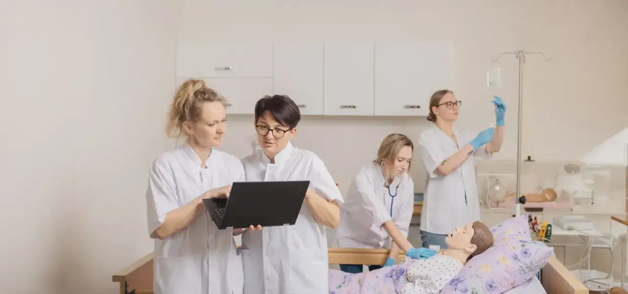 Pielęgniarka w białym kitlu uczy się obsługi laptopa, podczas gdy koleżanka ćwiczy zakładanie kroplówki przy fantomie.