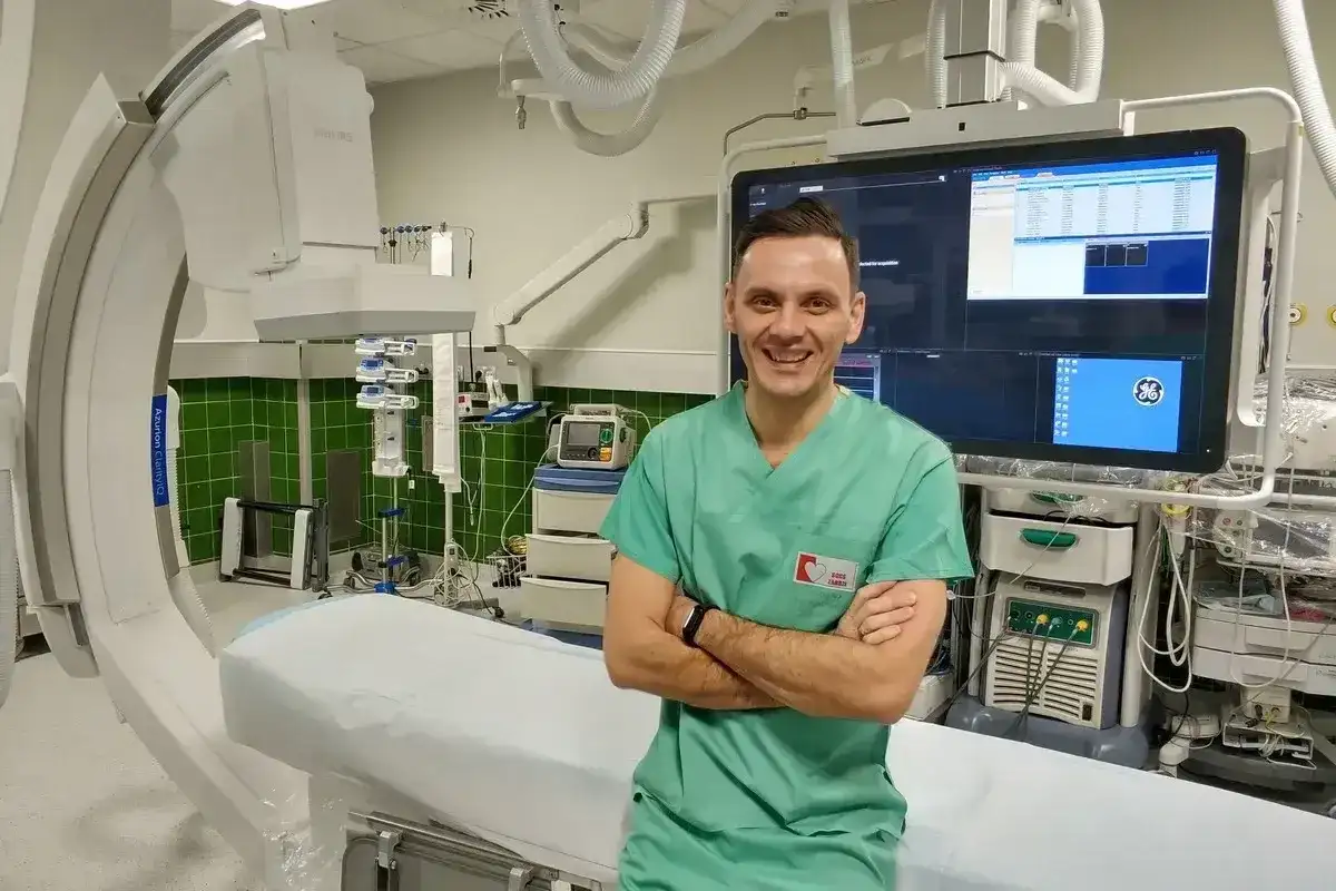 Uśmiechnięty technik elektroradiolog w zielonym fartuchu stoi na tle nowoczesnego sprzętu medycznego w sali zabiegowej.