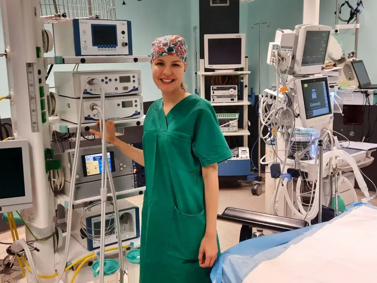 Uśmiechnięta instrumentariuszka w zielonym uniformie stoi obok aparatury medycznej na sali operacyjnej.