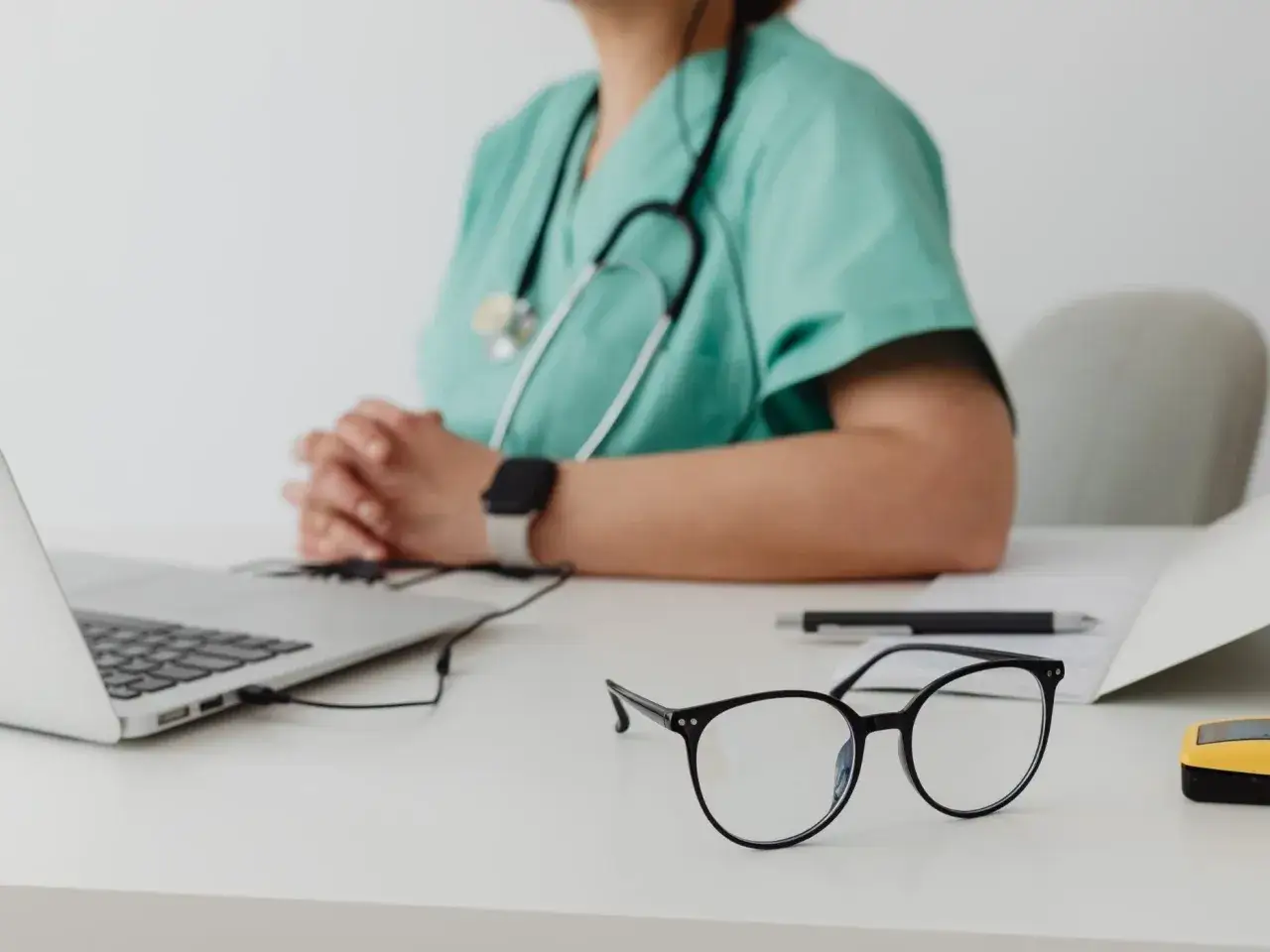 Lekarz w zielonym fartuchu zamyślony nad dokumentacją. Na biurku laptop, okulary i długopis. Czy to początek diagnozy choroby zawodowej?