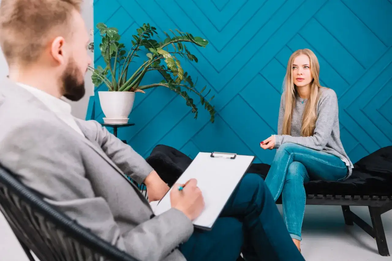 Rozmowa z psychologiem. Czy do psychiatry potrzebne jest skierowanie? Kobieta w szarej bluzie i jeansach siedzi naprzeciwko mężczyzny z notatnikiem.