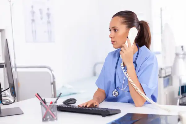Rejestratorka medyczna w niebieskim uniformie rozmawia przez telefon, pracując przy komputerze w gabinecie.