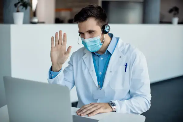 Lekarz w maseczce i kitlu wita pacjenta podczas konsultacji telemedycznej.