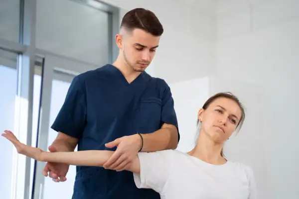 Fizjoterapeuta w granatowym uniformie delikatnie naciska na ramię pacjentki podczas ćwiczeń. Czy fizjoterapeuta to lekarz? Pomaga w rehabilitacji.