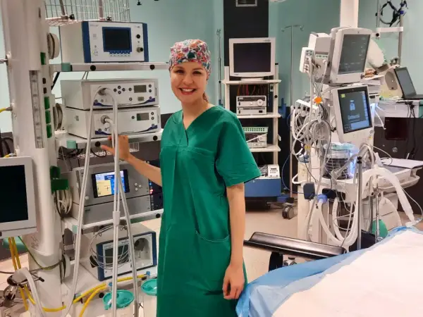 Uśmiechnięta instrumentariuszka w zielonym uniformie stoi obok aparatury medycznej na sali operacyjnej.